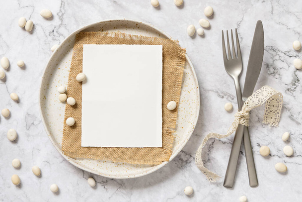 Пустая бумажная карточка, лежащая на белой тарелке с вилкой и ножом, завязанная винтажными лентами на мраморном столе с маленькими камнями вокруг, вид сверху. Карточный макет - Фото, изображение