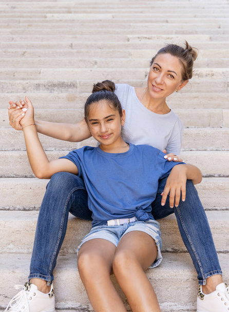 Mother and daughter sitting on stone stairs outdoor wearing t-shirts. A middle-aged woman holds hands of her kid girl . - Photo, Image