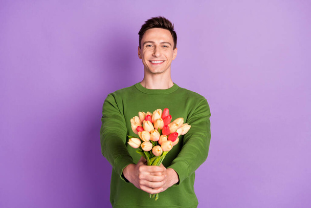 Photo of young flirty brunet guy give flowers wear sweater isolated on purple background - Zdjęcie, obraz