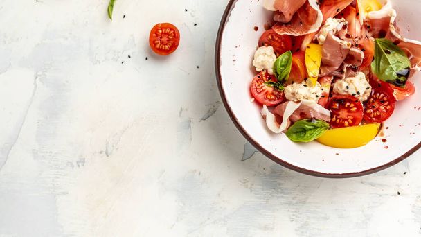 White bowl of fresh and healthy Mediterranean salad with tomatoes, cream cheese balls, prosciutto jamon and basil leaves. Delicious balanced food concept. Long banner format. top view. - Foto, imagen