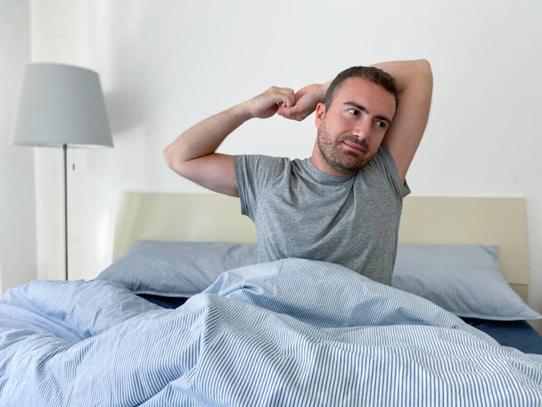 Ένας άντρας τεντώνει τους μυς και ξυπνάει από το κρεβάτι. - Φωτογραφία, εικόνα
