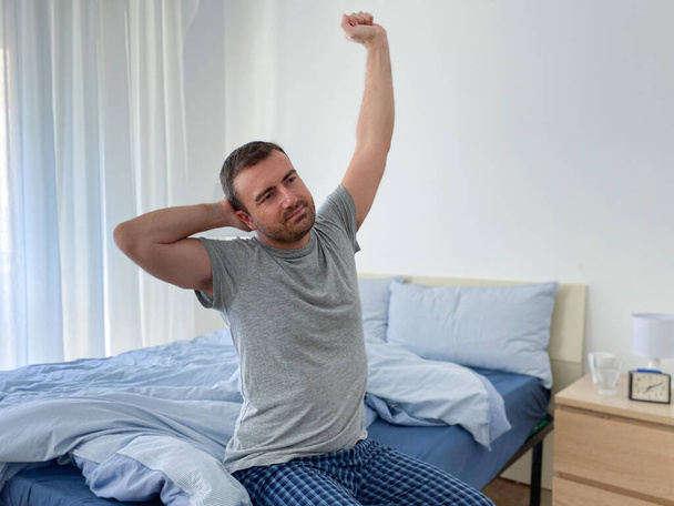 Ένας άντρας τεντώνει τους μυς και ξυπνάει από το κρεβάτι. - Φωτογραφία, εικόνα
