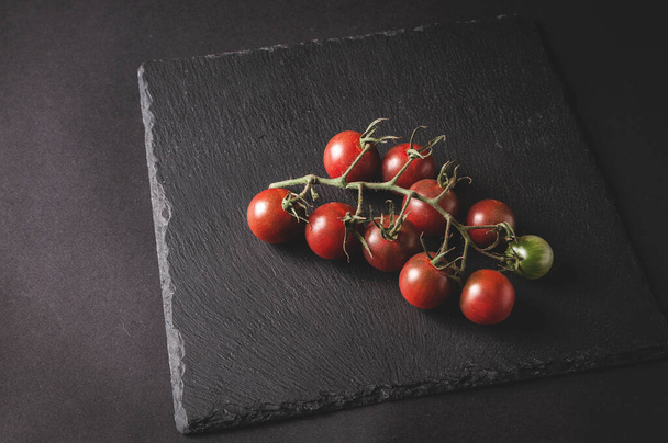 Ramo de tomates cherry frescos en una rama se encuentran en un tablero de servir negro sobre un fondo oscuro - Foto, imagen