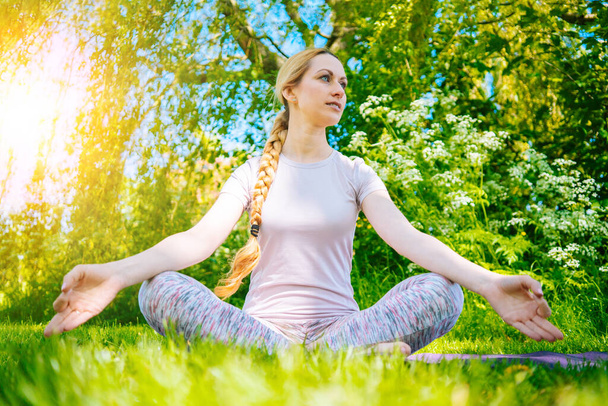 young woman doing yoga asana in park. girl stretching exercise in yoga position. happy and healthy woman sitting in lotus position and practicing yoga. meditation and sport on sunset outdoors. - Photo, Image