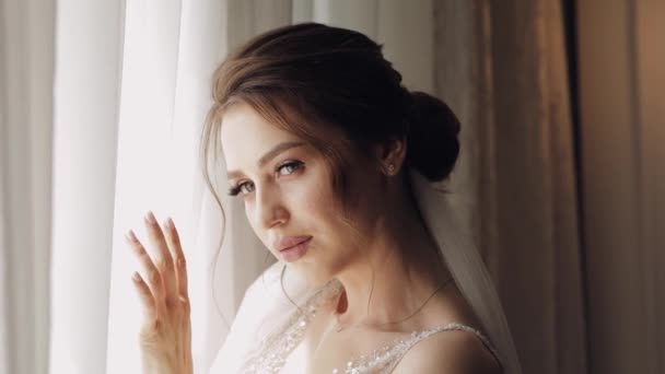 Bride in white dress staying near window and waiting for groom, wedding morning preparations at home - Footage, Video