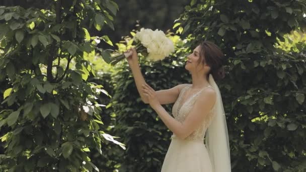 Krásná stylová nevěsta v bílých svatebních šatech a závoj drží svatební kytice v rukou v parku - Záběry, video