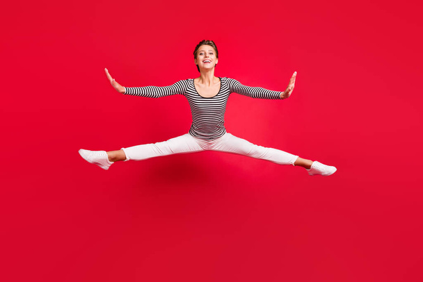 Photo of pretty cute young lady wear striped clothes smiling jumping high legs arms sides isolated red color background - Photo, Image