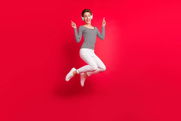 Photo of pretty sweet woman dressed striped outfit earphones jumping pointing fingers empty space isolated red color background - Foto, Imagem