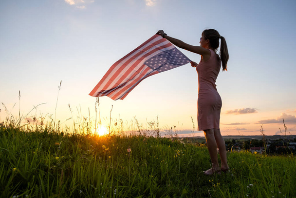Вид на счастливую молодую женщину, позирующую с национальным флагом США, стоящую на открытом воздухе на закате. Позитивная девушка празднует День независимости США. Концепция Международного дня демократии. - Фото, изображение