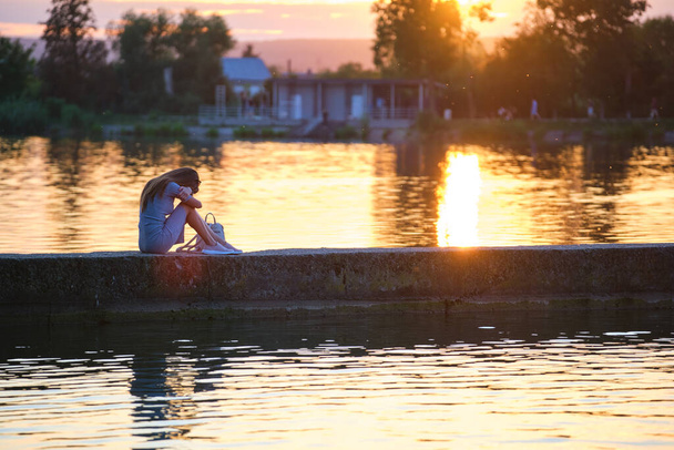 孤独な女性の側のビューだけで湖の海岸に暖かい夜に座っている。自然の中での孤独とリラックスの概念. - 写真・画像