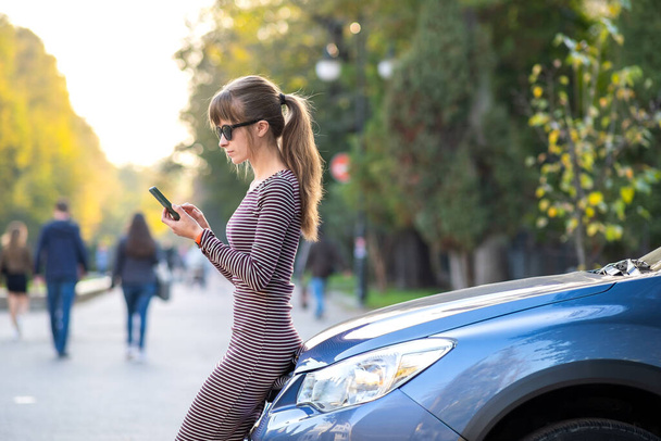 Νεαρή γυναίκα οδηγός στέκεται κοντά στο αυτοκίνητό της μιλώντας στο κινητό τηλέφωνο σε ένα δρόμο της πόλης το καλοκαίρι. - Φωτογραφία, εικόνα