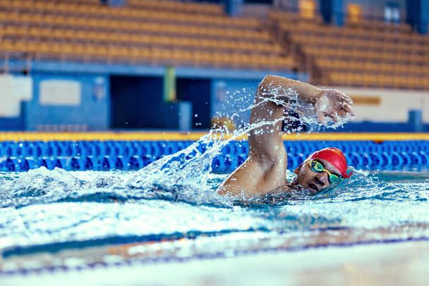 Professional male swimmer in swimming cap and goggles in motion and action during training at pool, indoors. Healthy lifestyle, power, energy, sports movement concept - Foto, afbeelding
