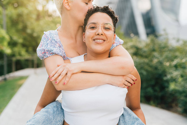 Two young multiethnic women hugging outdoor riding piggyback smiling joyful - 写真・画像