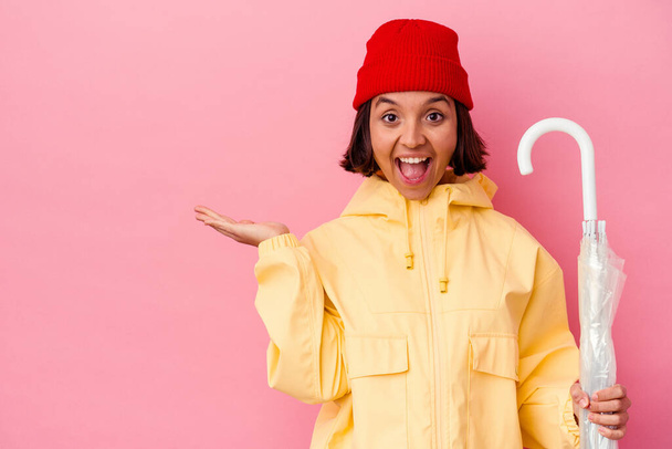 Young mixed race woman holding an umbrella isolated on pink background showing a copy space on a palm and holding another hand on waist. - Photo, Image