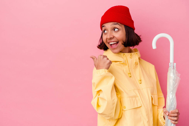 Young mixed race woman holding an umbrella isolated on pink background points with thumb finger away, laughing and carefree. - Photo, Image