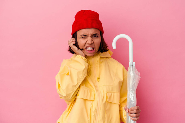 Young mixed race woman holding an umbrella isolated on pink background covering ears with hands. - Photo, Image