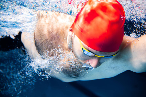 Close-up. Underwater view of professional male swimmer in red cap and goggles in motion and action during training at pool, indoors. Healthy lifestyle, power, energy, sports movement concept - Photo, image