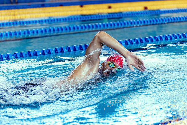 Professional male swimmer in swimming cap and goggles in motion and action during training at pool, indoors. Healthy lifestyle, power, energy, sports movement concept - Fotoğraf, Görsel