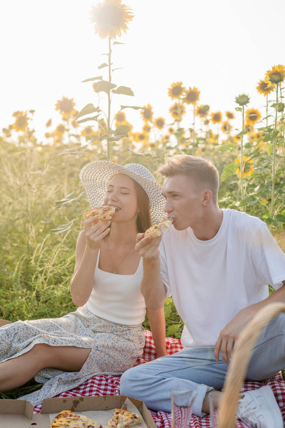 日没のひまわり畑で若い十代のカップルピクニック。ピザやシャンパンを食べる - 写真・画像