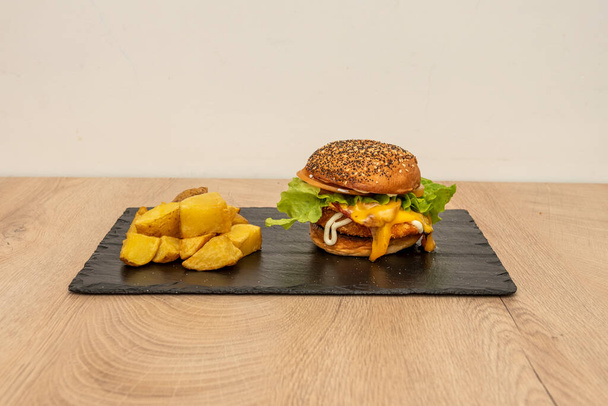 Klassischer knuspriger Chicken Burger mit geschmolzenem Cheddar-Käse, gebratenem Speck, Mayonnaise, Senf- und Tomatenscheiben mit Salat und gehackten Bratkartoffeln garniert - Foto, Bild