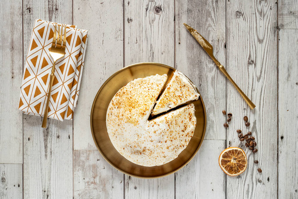 Top view image of carrot cake with frosty bench, golden plates and cutlery on white wooden table with coffee beans and dried orange - Foto, Imagen