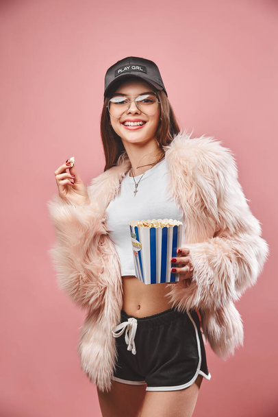 Attractive brunette girl in faux pink fur holding popcorn in hand. Wearing in black shorts white top black cap and glasses. Looking at the camera. Studio pink background - Photo, image