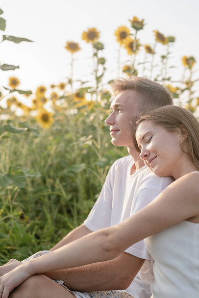 Herbstliche Natur. Spaß und Lebensfreude. Junges Teenie-Paar picknickt auf Sonnenblumenfeld im Sonnenuntergang, hat Spaß beim Anblick einer Frau, die auf der Schulter ihres Freundes liegt - Foto, Bild