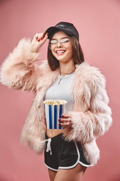Attractive brunette girl in faux pink fur holding popcorn in hand. Wearing in black shorts white top black cap and glasses. Looking at the camera. Studio pink background - Foto, Bild