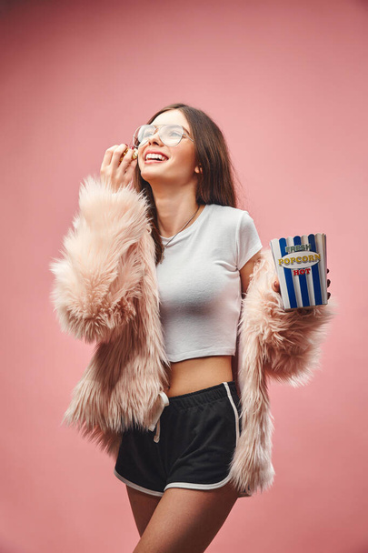 Laughing brunette girl in faux pink fur holding popcorn in hand. Wearing in black shorts white top and glasses. Looking up. Studio pink background. Girl with long dark hair - Fotó, kép