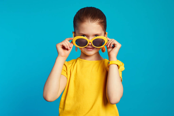 Κορίτσι με κίτρινο πουκάμισο ρυθμίζοντας γυαλιά ηλίου και κοιτάζοντας κάμερα μέσα από αυτά - Φωτογραφία, εικόνα