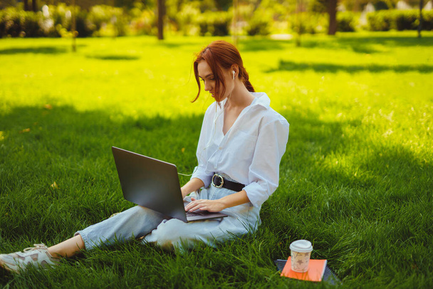 Beautiful young pretty redhead woman in park outdoors using laptop computer for study or work, wired headphones - Photo, Image
