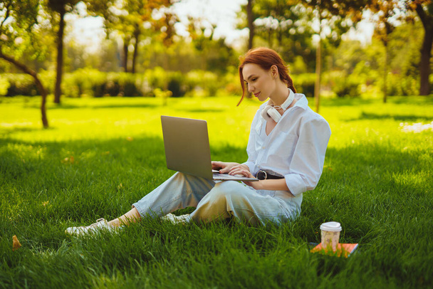 Beautiful young pretty redhead woman in park outdoors using laptop computer for study or work online, wireless earphones on her neck - Foto, Imagem