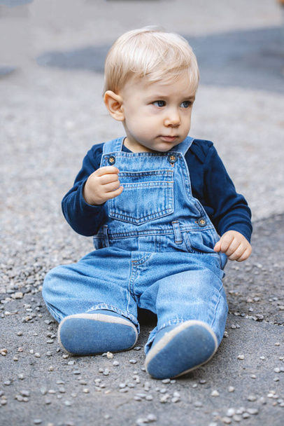 Mała blondyneczka siedząca na ziemi. Chłopiec w niebieskich dżinsach. - Zdjęcie, obraz