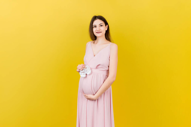 Счастливая беременная женщина держит маленькие девчачьи ботинки на животе на цветном фоне. Счастливой беременности. Копирование пространства. - Фото, изображение