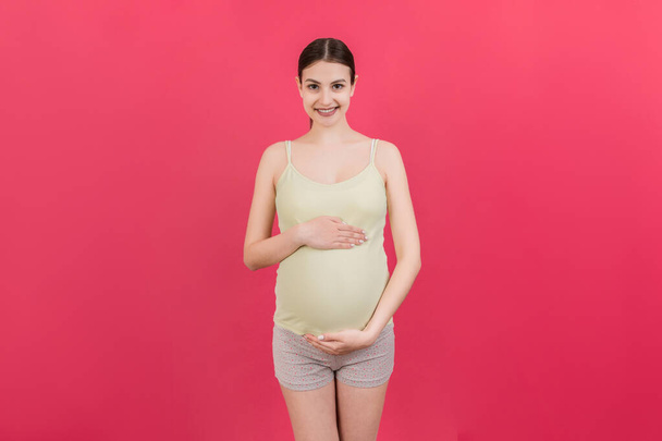Happy pregnant woman touching her abdomen at Colored background. Future mother is wearing white underwear. Expecting of a baby. Copy space. - Photo, Image