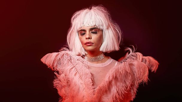 Tired drag queen, young transgender with rubbed bright night makeup wearing boa feather party dress and white wig, posing isolated in red neon light background - Photo, Image