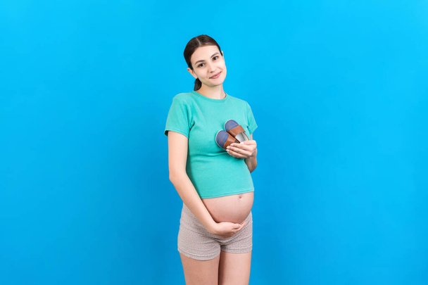Счастливая беременная женщина показывает маленькие сапоги для мальчика на цветном фоне. Будущая мать ждет ребенка. Копирование пространства. - Фото, изображение