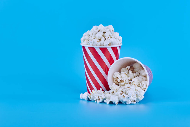 Dvě červené sklenice popcornu na modrém pozadí, jedna z nich leží a z ní vytéká popcorn - Fotografie, Obrázek