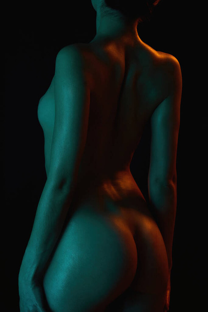 Back view of naked body of sexy young woman standing and posing isolated in trendy neon light over dark background - Фото, зображення