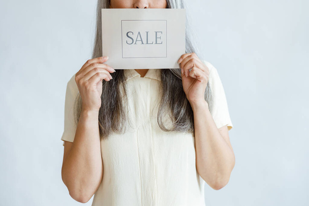 Mature woman with long grey hair holds Sale sign standing in studio closeup - Fotó, kép