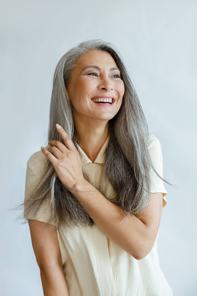 Middle aged Asian woman touches natural hoary hair on light grey background - Zdjęcie, obraz