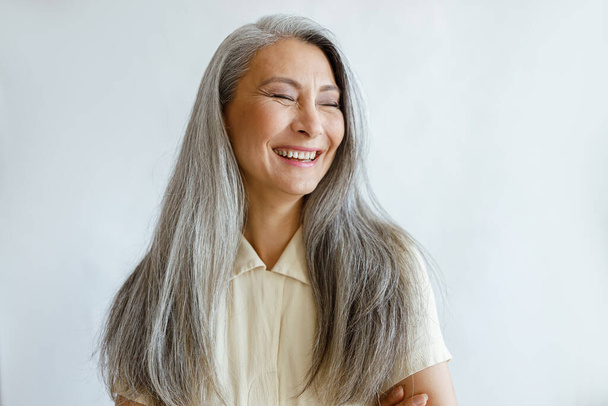 Портрет улыбающейся азиатки с седыми волосами и скрещенными руками на сером фоне - Фото, изображение