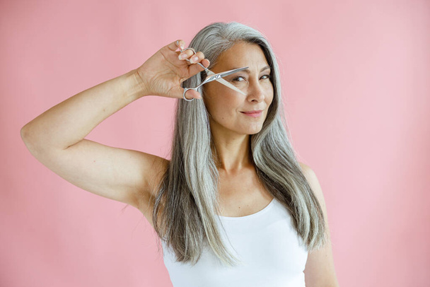 Belle dame asiatique aux cheveux gris tient ciseaux ouverts près du visage sur fond rose - Photo, image