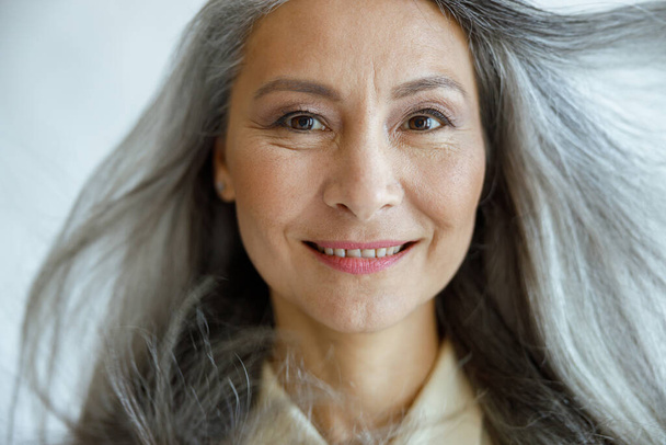 Middle aged Asian woman with flying hoary hair looks into camera on light grey background - Photo, Image