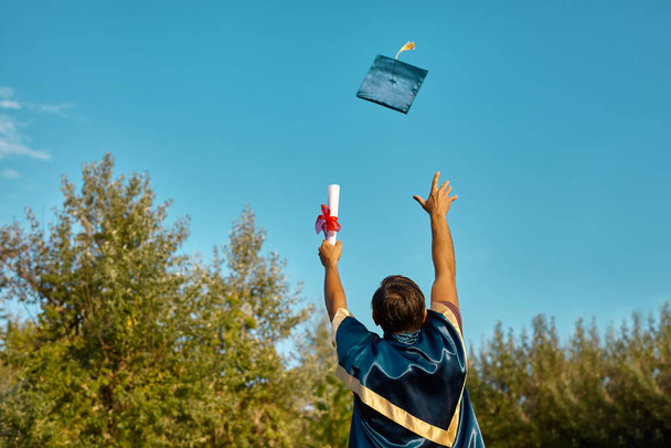 vzrušený postgraduální student v róbě s vystouplýma rukama držící diplom nad modrou oblohou a hází do nebe klobouk - Fotografie, Obrázek