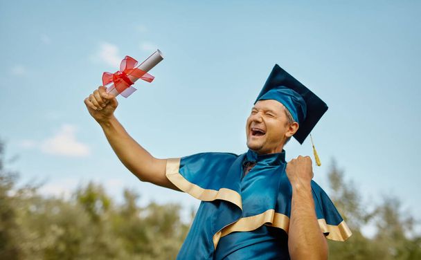 Mladý šťastný usměvavý muž slaví svůj diplom a diplom po modré obloze. oslava konceptu promoce - Fotografie, Obrázek