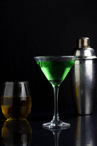 A vertical shot of a vibrant green drink on a dark black background with a shaker and glass behind - Φωτογραφία, εικόνα