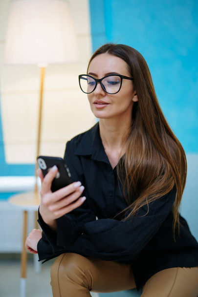 Stunning beautiful girl smiling and poses to the camera in modern glasses. Woman sitting and looking at the smartphone. Light interior colors. Closeup - Foto, imagen