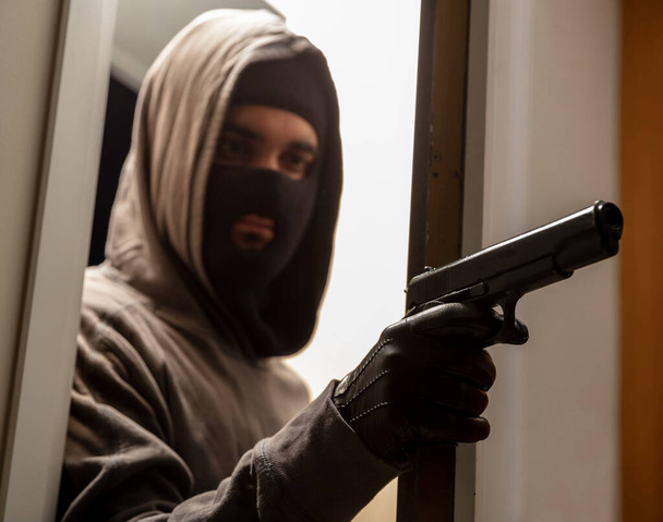 Ready for invasion concept. Thief robber killer burglar gangster terrorist armed young man in grey hoodie with black balaclava holding pistol in leather glove behind half opened door. Target attack. - Foto, afbeelding