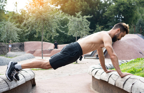 Outdoor fitness. Young athletic bearded man doing push up exercise using objects of the urban environment.  - Zdjęcie, obraz